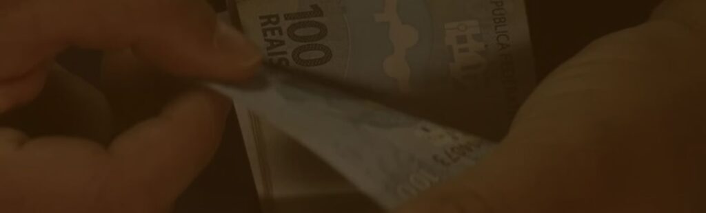 A imagem mostra uma pessoa manuseando cédulas de dinheiro, especificamente notas de 100 reais, sugerindo um foco em transações financeiras ou tributárias. A foto captura o momento em que uma nota é retirada de um maço, destacando a ação de contar ou distribuir dinheiro. Essa imagem pode estar relacionada a discussões sobre impostos, finanças pessoais ou temas econômicos no contexto da Reforma Tributária.